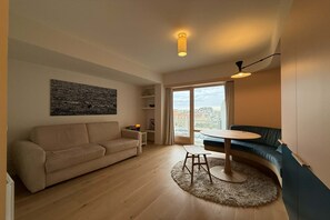 Ein Wohnzimmer mit einer Couch, einem kleinen Tisch, einem runden Teppich, einer Deckenleuchte und einem großen Fenster mit Vorhängen.
