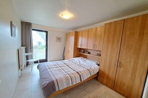 Quarto com cama, guarda-roupa de madeira, radiador, cadeira e janela com cortinas.