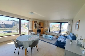 Ampla sala de estar com mesa de jantar, sofá e janelas grandes que oferecem vista para o exterior.