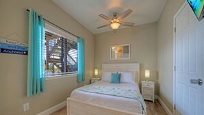 Ein Schlafzimmer mit einem Bett, einem Deckenventilator, einem Fenster mit Vorhängen, einem Nachttisch, einer Lampe und einer Tür.