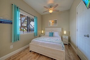 Ein Schlafzimmer mit einem Bett, einem Deckenventilator, einem Fenster mit Vorhängen, einem Nachttisch, einer Lampe und einer Tür.