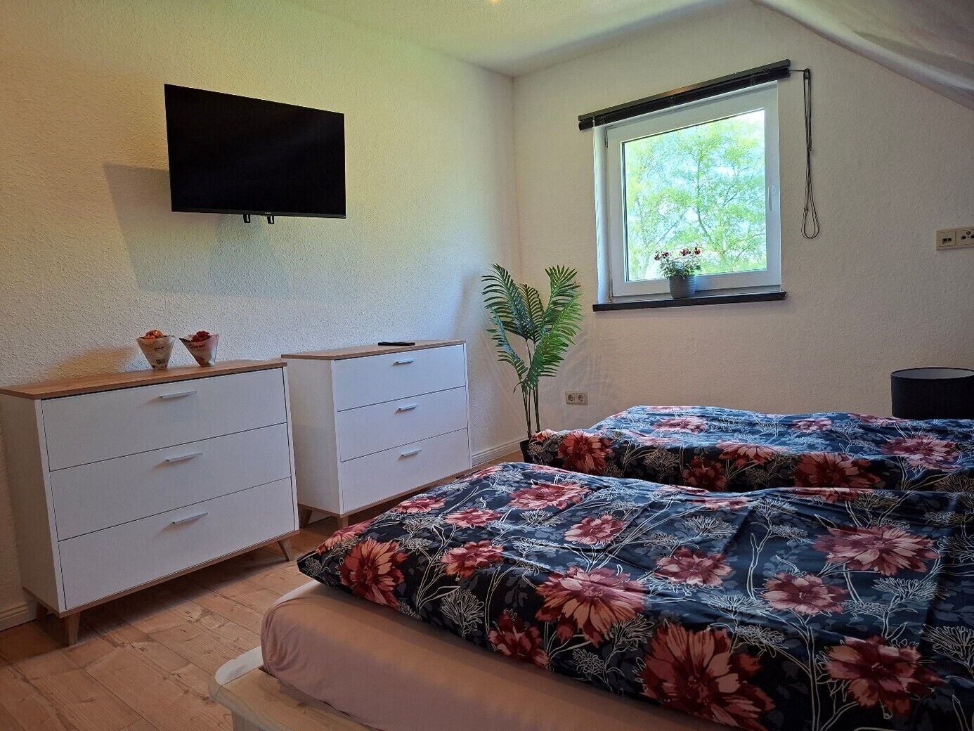A bedroom with a bed, a TV mounted on the wall, a window with a view of greenery, and a plant beside the window.