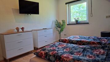 A bedroom with a bed, a TV mounted on the wall, a window with a view of greenery, and a plant beside the window.