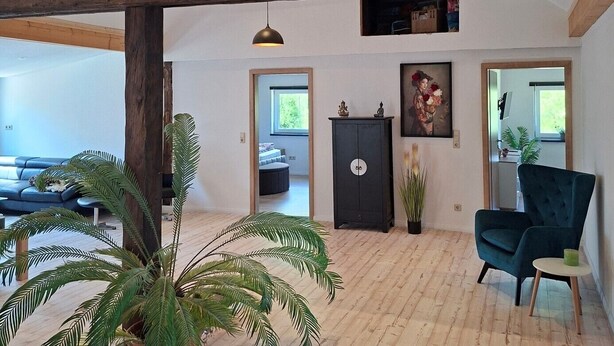 A spacious room with wooden beams, a sofa, a chair, and potted plants.