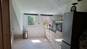 A kitchen with a wooden floor, a stainless steel refrigerator, and a window with a view of trees.