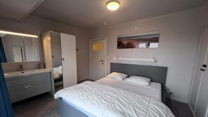 Ein Schlafzimmer mit Bett, Kommode, Spiegel, einer Tür und einer Wandleuchte.