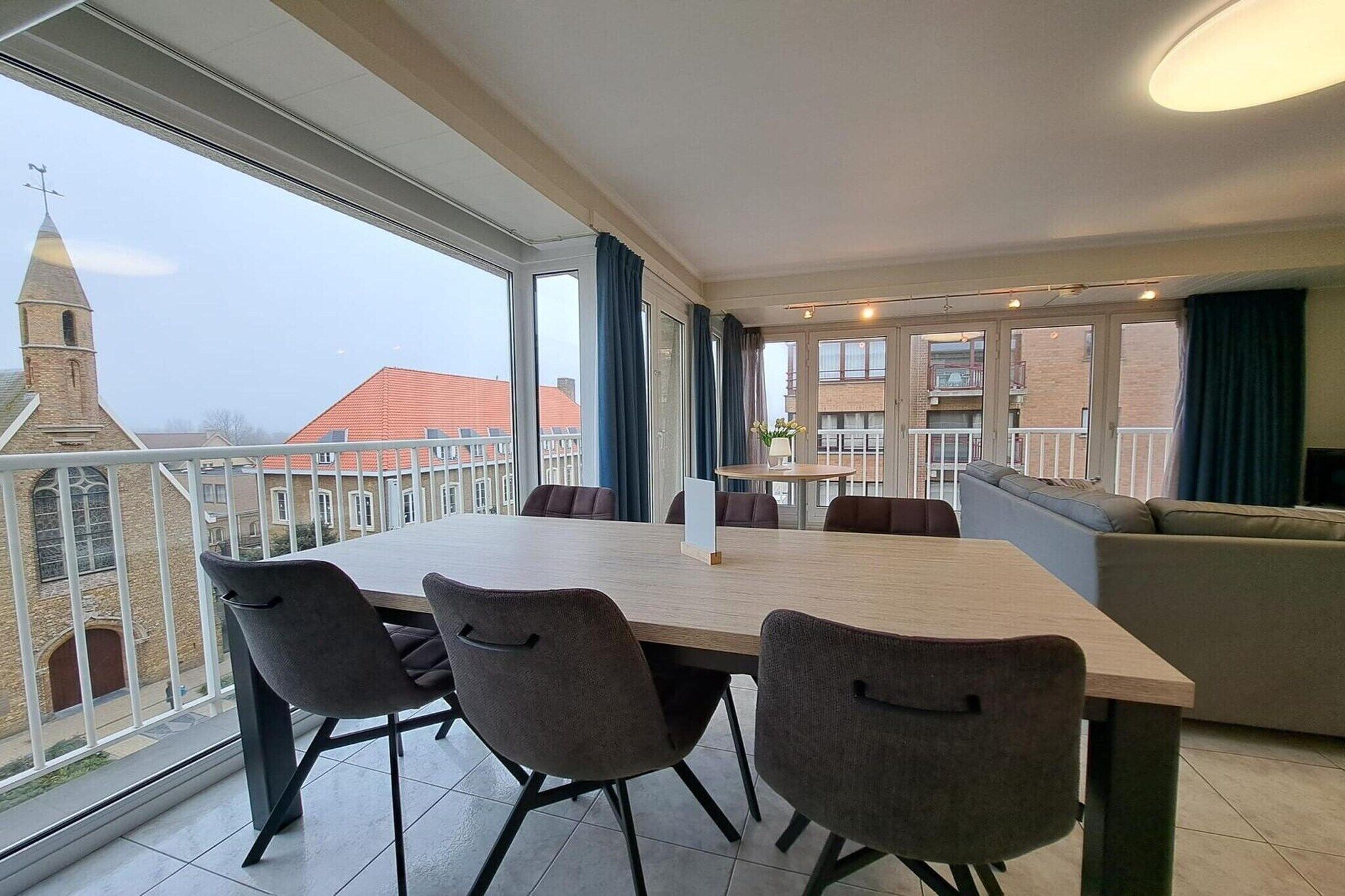 Una habitación moderna con una mesa grande y sillas, un sofá, y un balcón que ofrece vistas a edificios y una torre.