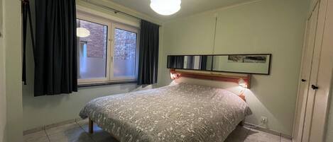 Un dormitorio con cama, cortinas en la ventana, lámpara de noche y un cuadro colgado en la pared.
