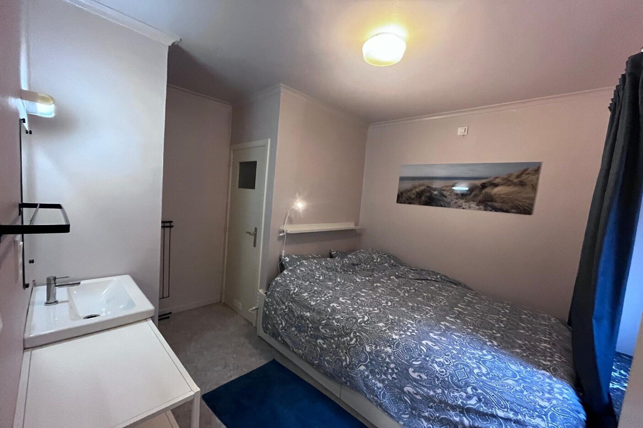 A small, single-bed room with a white sink, a mirror, a door, a bed with a patterned bedspread, a wall-mounted shelf, and a picture.