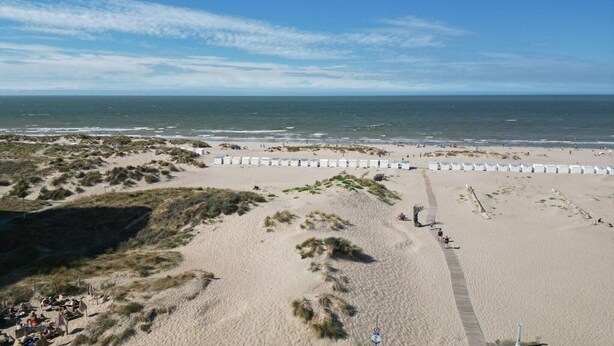 Ein Strand mit einem Sandweg, der zu einer weißen Umzäunung führt, wolkenloser Himmel und dem Meer im Hintergrund.