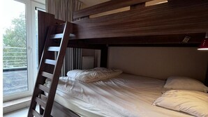Een stapelbed met een houten trap, een raam met gordijnen en een radiator.