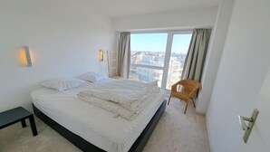 Quarto com cama, cadeira, mesa de cabeceira e vista da cidade através de janelas amplas.