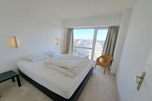 Ein Schlafzimmer mit Bett, Stuhl, Nachttisch und Blick auf die Stadt durch große Fenster.