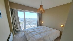 Quarto com cama grande, uma cadeira, luminária pendente e vista para a cidade através de uma janela.