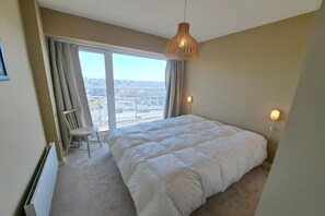 Ein Schlafzimmer mit einem großen Bett, einem Sessel, einer Hängelampe und Blick auf eine Stadtansicht durch ein Fenster.