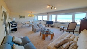 Uma sala de estar espaçosa com uma grande janela com vista para a praia, uma televisão e um espaço para refeições.