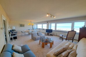 Ein geräumiges Wohnzimmer mit großem Fenster zum Strand, einem Fernseher und einem Essbereich.