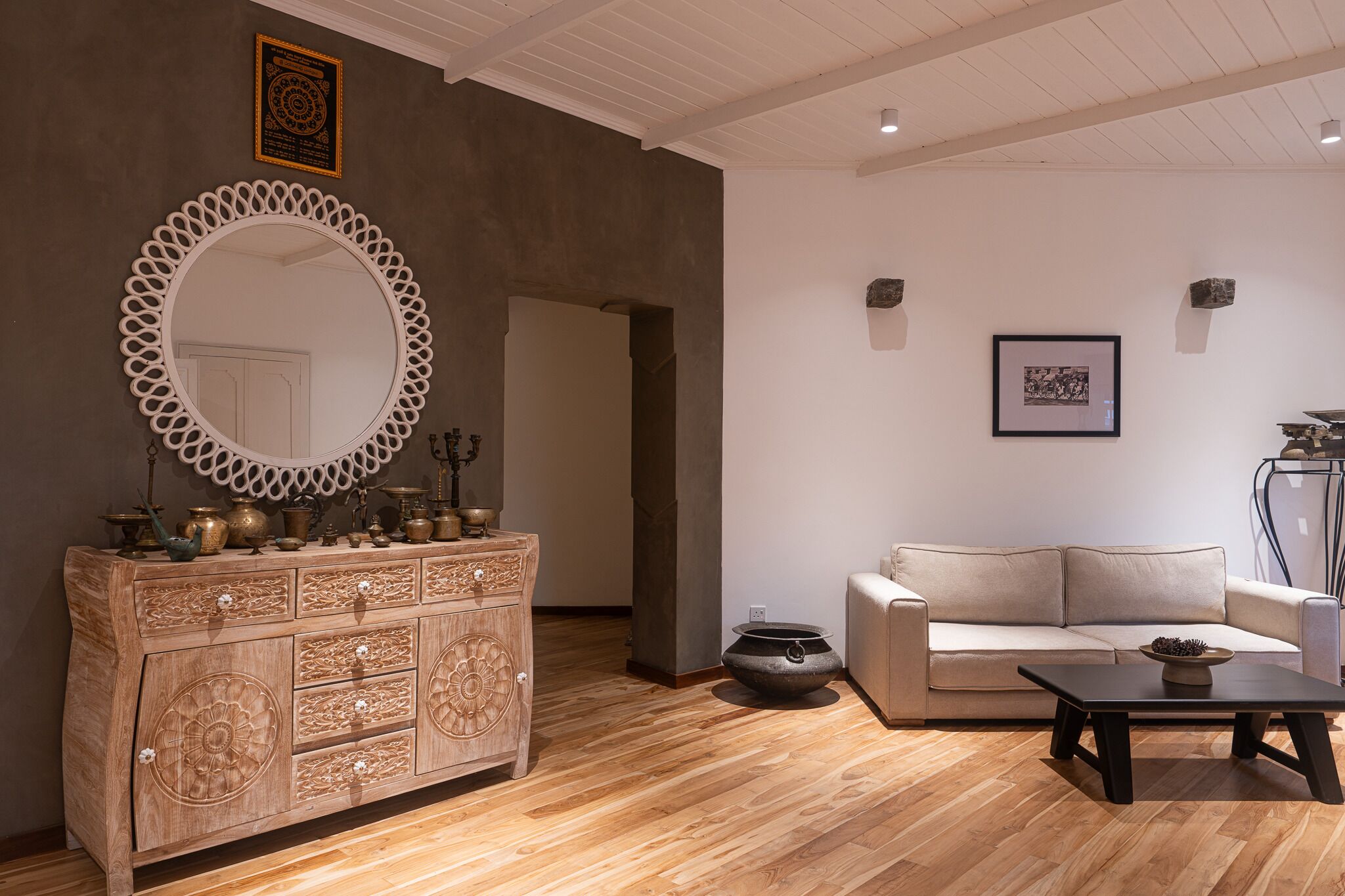 Una sala de estar moderna con piso de madera, un sofá color beige, una mesa de centro negra y un mueble de madera decorativo con objetos de latón.