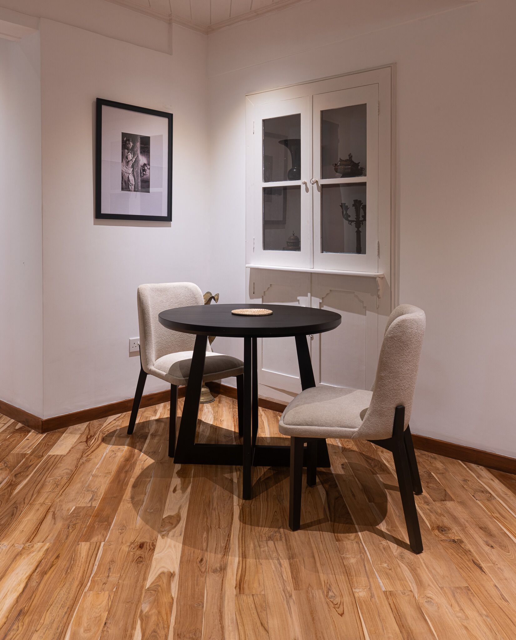 Una habitación con una mesa redonda y dos sillas, un cuadro enmarcado en la pared, y un aparador con puertas de vidrio.