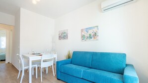 Ein Zimmer mit einem blauen Sofa, einem weißen Esstisch mit Stühlen und zwei an der Wand hängenden Bildern.