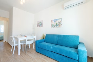 Ein Zimmer mit einem blauen Sofa, einem weißen Esstisch mit Stühlen und zwei an der Wand hängenden Bildern.