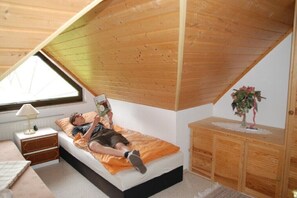 Ein Schlafzimmer mit schrägem Dach, einem Bett mit orangefarbener Decke, einem Nachttisch mit Lampe und einem Holzschrank.
