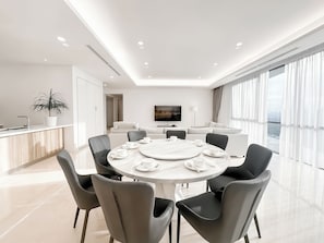 Una habitación de hotel moderna con zona de comedor, sofá, televisor y un ventanal grande.