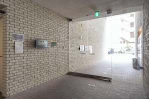 A brick-walled corridor with a glass door leading to an outdoor area, a green exit sign, and a fire alarm panel. - T-FLAT NAMBA WEST (Sakuragawa, Naniwa)