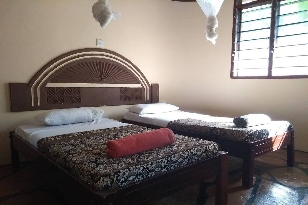 Una habitación con dos camas, una colcha estampada, un cojín rojo y una ventana con persianas.