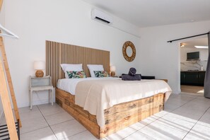A bedroom with a wooden bed frame, a bedside table with a lamp, a wall-mounted TV, and a kitchen area visible through an open door. - Villa "De L’Olivier" with Private Pool, Wi-Fi, and Air Conditioning (Labégude)