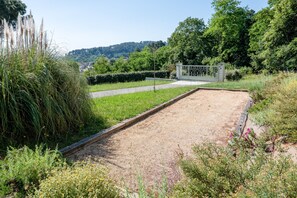 Un jardín con una cancha de arena, un portón metálico y vistas a una colina con árboles.