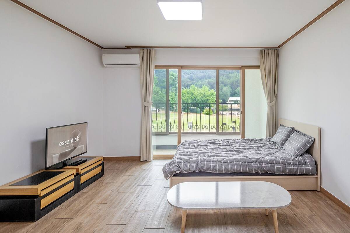 Un dormitorio con cama, televisor, escritorio y balcón con vistas a la vegetación.