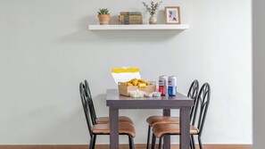 Una mesa de comedor con sillas, una caja de pollo frito, dos latas de refresco y un pequeño jarrón con una planta en un estante superior.