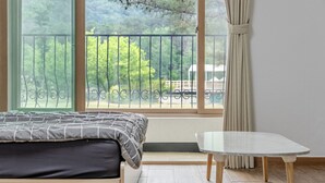 A bedroom with a bed, a small table, a balcony with a railing, and a view of trees and a building. - Chuncheon O2 Pension (Chuncheon)