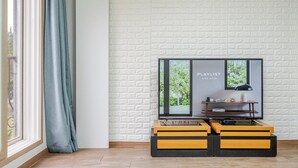 A room with a white brick wall, a large window with a view of greenery, a television on a wooden stand, and a black lamp. - Chuncheon O2 Pension (Chuncheon)