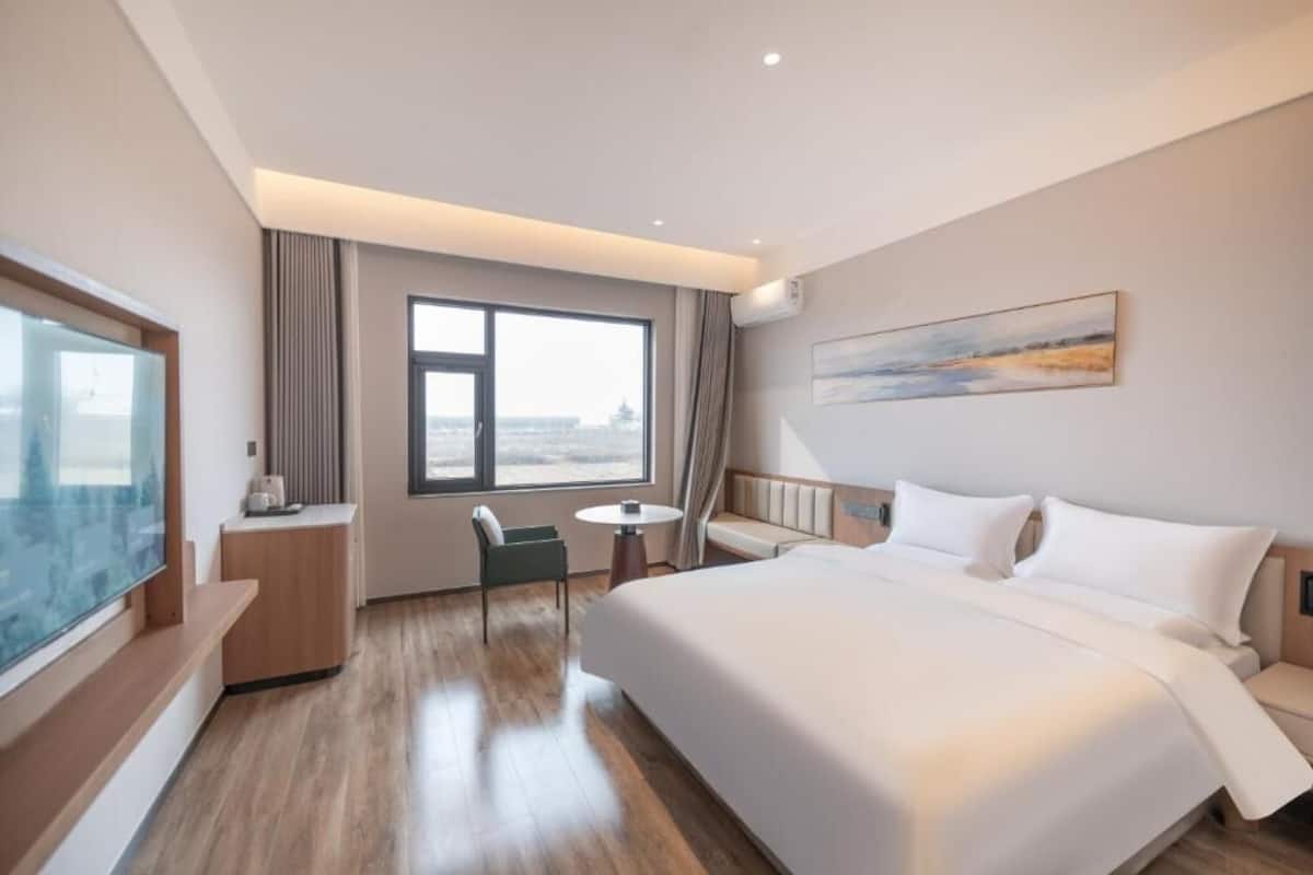 A hotel room with a large bed, a seating area with a chair and a small table, a television, and a large window.