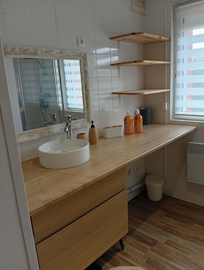 Une salle de bain moderne avec un lavabo blanc, un meuble-lavabo en bois et un miroir doté d’un cadre orné. On trouve des étagères en bois au-dessus du lavabo et une fenêtre avec des stores.