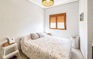 Ein Schlafzimmer mit einem Bett, einem Nachttisch, einer Lampe, einem Fenster mit Jalousien und einer Deckenleuchte.