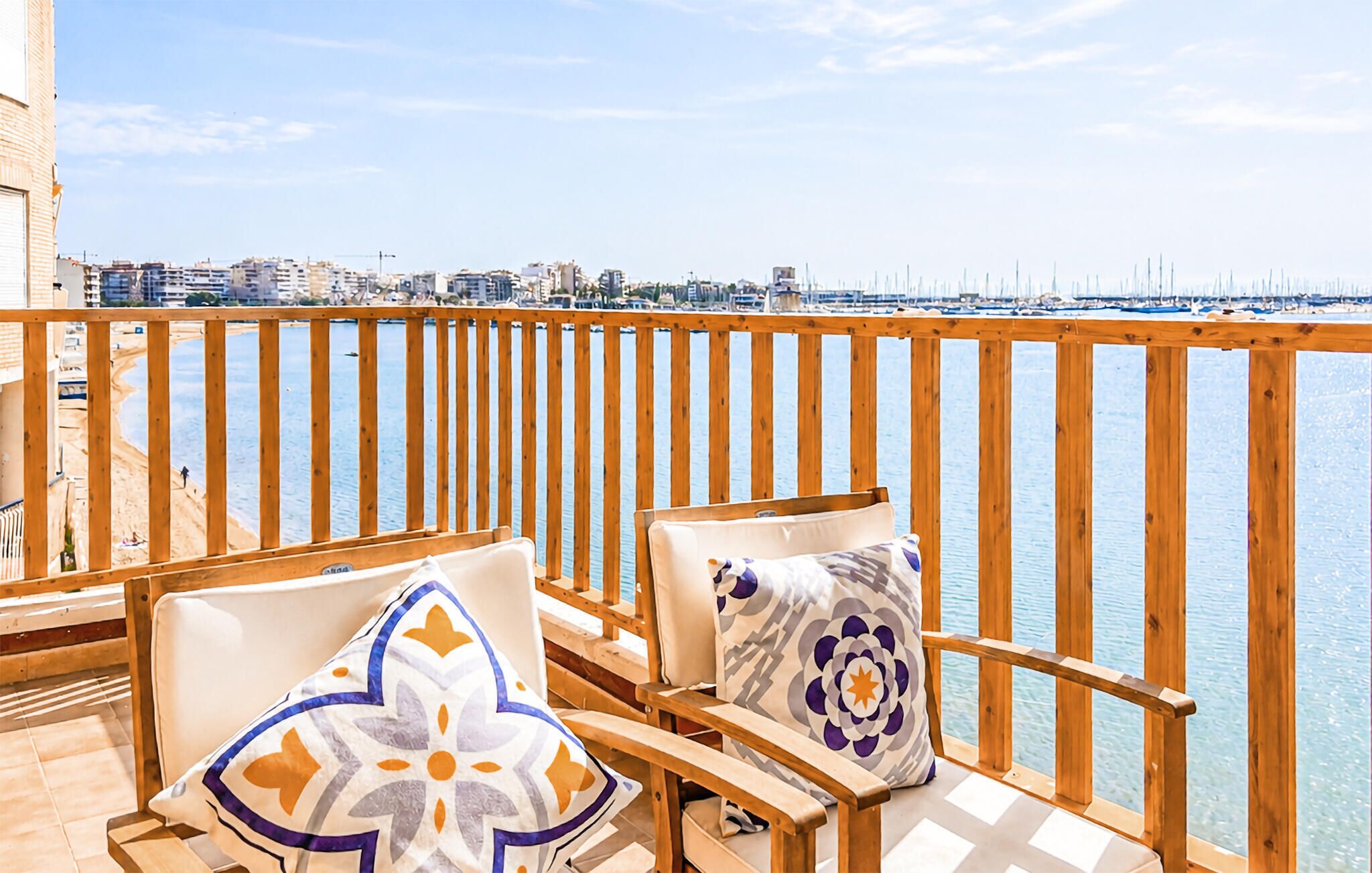 Un balcón con barandillas de madera, dos sillas con cojines y vista a un puerto deportivo y paisaje urbano.