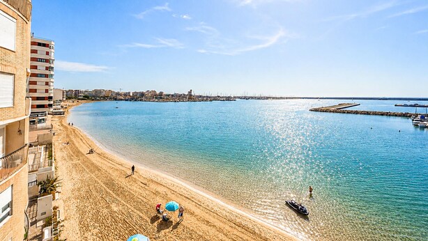 A coastal beach with clear water, a pier, and a building with balconies. - 2 bedroom beach front apartment in Torrevieja (Torrevieja)