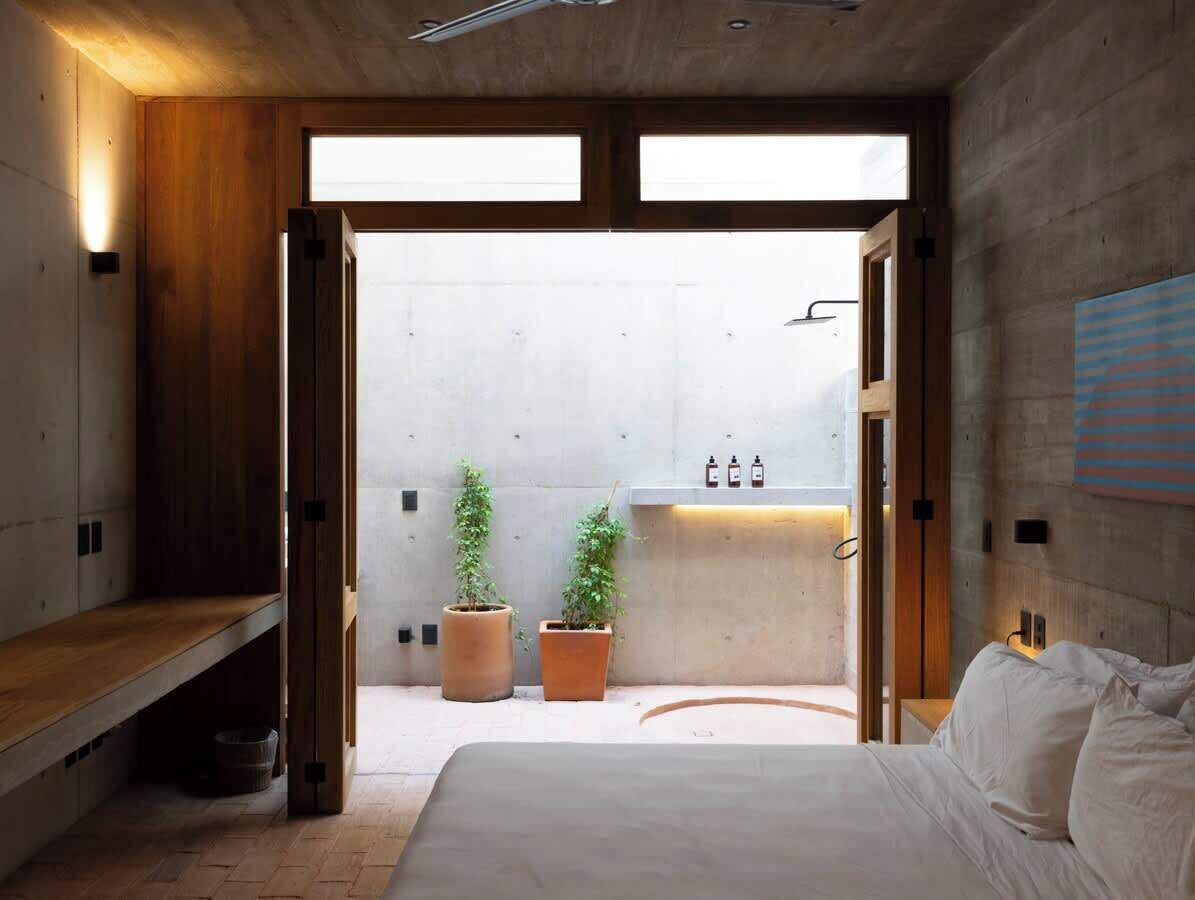 Une chambre à coucher moderne avec un lit, un banc en bois, des plantes en pot et une salle de bain équipée d’un lavabo et d’une douche.