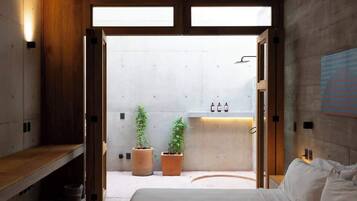 Une chambre à coucher moderne avec un lit, un banc en bois, des plantes en pot et une salle de bain équipée d’un lavabo et d’une douche.