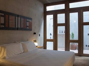 A bedroom with a large bed, a nightstand, a wall-mounted light, and a view through large windows to an outdoor area with a potted plant. - 2BR Architectural Retreat with Private Pool NEW (Brisas de Zicatela)