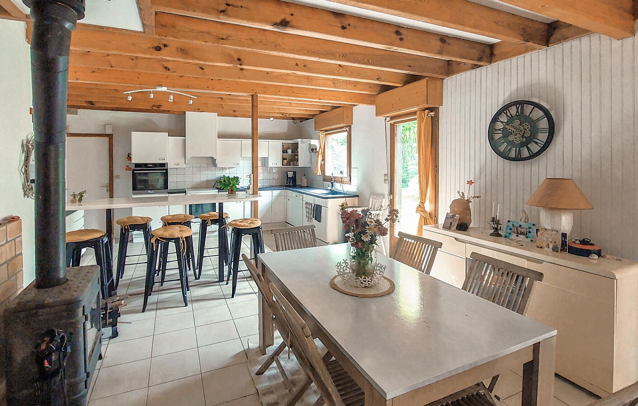A kitchen with wooden beams, a dining area with stools, a clock on the wall, and a window with curtains.