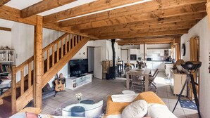 Un espacio habitable amplio con una escalera de madera, televisión, mesa de comedor y cocina al fondo.