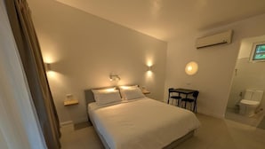 Un dormitorio con cama, mesitas de noche, una pequeña mesa, una silla, unidad de aire acondicionado, ventana y un inodoro.
