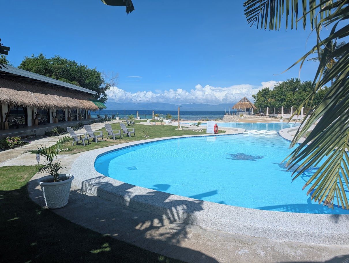 A resort pool with lounge chairs, a thatched-roof building, and a view of the ocean.