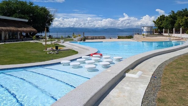 Una piscina con vistas al océano, rodeada por un área verde y un edificio con techo de paja.