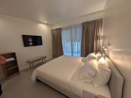 Quarto com cama grande, televisão de tela plana na parede, um banquinho de madeira e janela com cortinas.