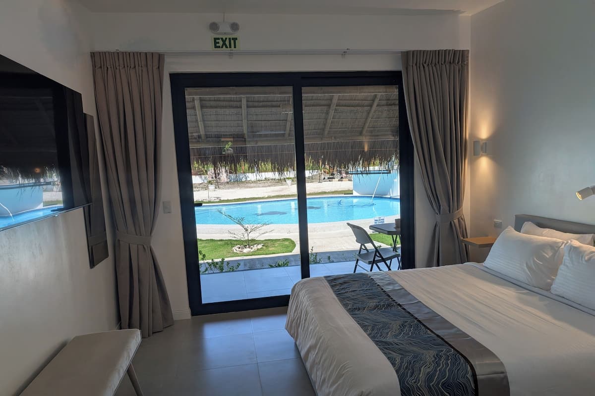 A hotel room with a bed, a bench, a TV, a pool view, and an exit sign.